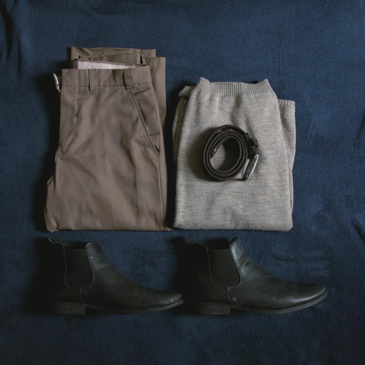 Men's clothing ensemble including trousers, sweater, boots, and belt arranged on a blue surface.